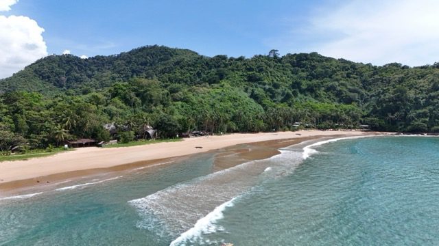 Aerial view of Duli Beach, with waves rolling onto sand backed by forested hills